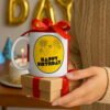 happy-birthday-coffee-mug-gift-idea Girt-ready White mug with 'Happy Birthday' design on a small box, held by a person with a cake and candles in the background.